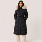 A woman wearing a long black puffer coat with a large beige fur-trimmed hood and a matching tie belt, paired with light beige pants, against a plain light beige studio background.
