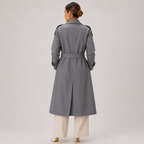 A woman viewed from the back wearing a long gray trench coat with a matching belt, paired with light beige wide-leg pants and beige heels, against a plain light gray studio background.