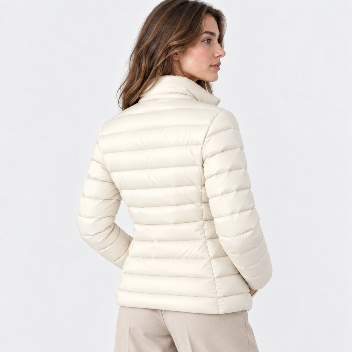 A woman shown from the back wearing a cream-colored, horizontally quilted puffer jacket and light beige trousers, set against a plain light gray studio background.