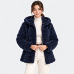 A woman modeling a navy blue sherpa fleece puffer jacket with a high collar, worn over a cream ribbed turtleneck and cream trousers, against a light gray studio background.
