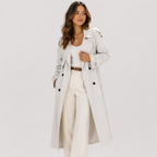 A woman modeling a long light gray double-breasted trench coat over a white top and white wide-leg trousers, photographed against a plain light gray studio background.