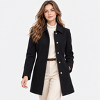 A woman modeling a black mid-length coat over a cream ribbed turtleneck and cream trousers, photographed against a clean white studio background.