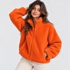 A woman wearing a bright orange sherpa fleece zip-up jacket and light beige pants, posing against a solid light gray background.