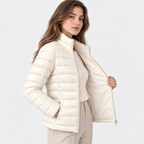 A woman modeling a cream-colored puffer jacket with a high stand-up collar, shown against a plain light gray studio background.