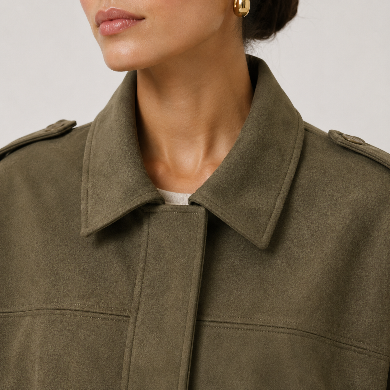 Close-up of a model wearing an olive green suede-like jacket with a structured collar and shoulder tab details, paired with a white top and a gold hoop earring.