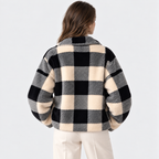 A woman viewed from behind wearing a fuzzy black and cream plaid jacket with wide sleeves, paired with cream pants, against a light gray studio background.