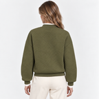 A woman viewed from the back wearing an olive green textured knit sweater with a diamond pattern, paired with cream-colored pants, against a plain white studio background.