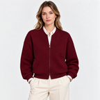 A woman wearing a textured burgundy zip-up bomber jacket over a white collared shirt and cream trousers, standing against a light gray studio background.