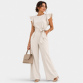 A woman models a cream-colored ruffled sleeve jumpsuit with a matching waist tie, paired with a beige handbag and nude heels, against a light gray studio background.