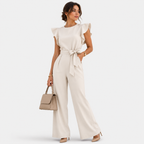 A woman models a cream-colored ruffled sleeve jumpsuit with a matching waist tie, paired with a beige handbag and nude heels, against a light gray studio background.