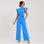 A woman modeling a bright blue wide-leg jumpsuit with ruffled cap sleeves and a matching waist tie, photographed against a plain light gray studio background.