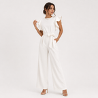 A woman modeling a white wide-leg jumpsuit with ruffled short sleeves and a matching waist tie, photographed against a clean white studio background.