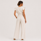 A woman shown from the back wearing a light cream jumpsuit with ruffled cap sleeves, a defined waist, and wide legs, set against a plain light gray studio background.