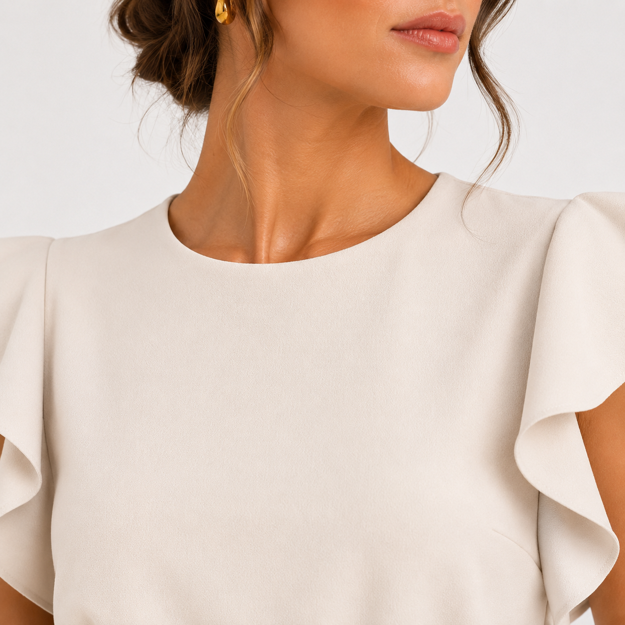 Close-up of a woman wearing a cream-colored blouse with ruffled short sleeves and a round neckline, set against a plain light background.