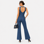 A woman modeling a blue sleeveless flared jumpsuit from the back, featuring a large open back design, paired with black high heels and a small black handbag, set against a plain light gray background.