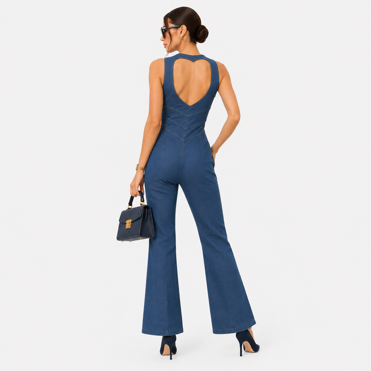 A woman modeling a blue sleeveless flared jumpsuit from the back, featuring a large open back design, paired with black high heels and a small black handbag, set against a plain light gray background.