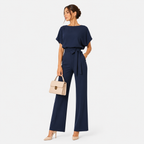 A woman modeling a navy blue two-piece jumpsuit with a tie waist and flutter sleeves, holding a light beige handbag against a plain white studio background.