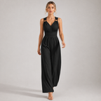 A woman modeling a black sleeveless jumpsuit with cutout shoulder details, a V-neckline, and wide-leg pants against a plain light gray studio background.
