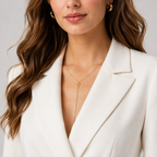 A close-up of a woman wearing a delicate gold Y-necklace with a small geometric pendant, styled with gold hoop earrings and a white blazer.