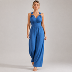 A woman modeling a sleeveless blue jumpsuit with a gathered waist and wide legs, standing against a plain light gray studio background.