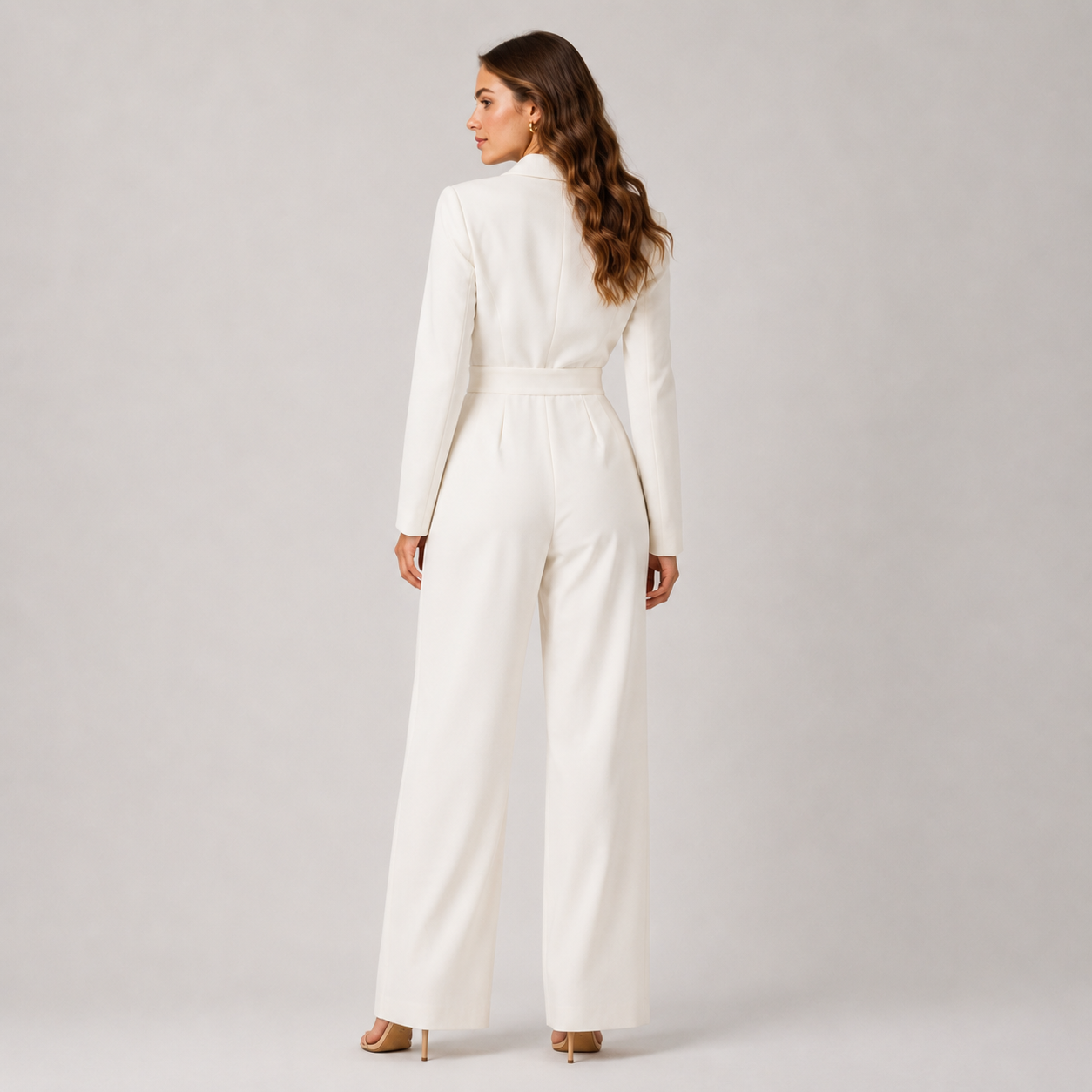 A woman viewed from the back wearing a white long-sleeve jumpsuit with a defined waist and wide legs, standing against a light gray studio background.