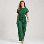 A woman modeling a green one-piece jumpsuit with a white polka dot pattern, featuring short flutter sleeves, a boat neckline, and a matching fabric tie belt at the waist, set against a plain light gray studio background.