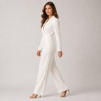 A woman modeling a white long-sleeve jumpsuit with a deep V-neckline, structured shoulders, and wide-leg trousers, paired with nude high-heeled sandals against a light gray studio background.