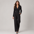 A woman modeling a black long-sleeve jumpsuit with a wrap-style V-neckline, defined waist, and wide legs, photographed against a neutral light gray studio background.