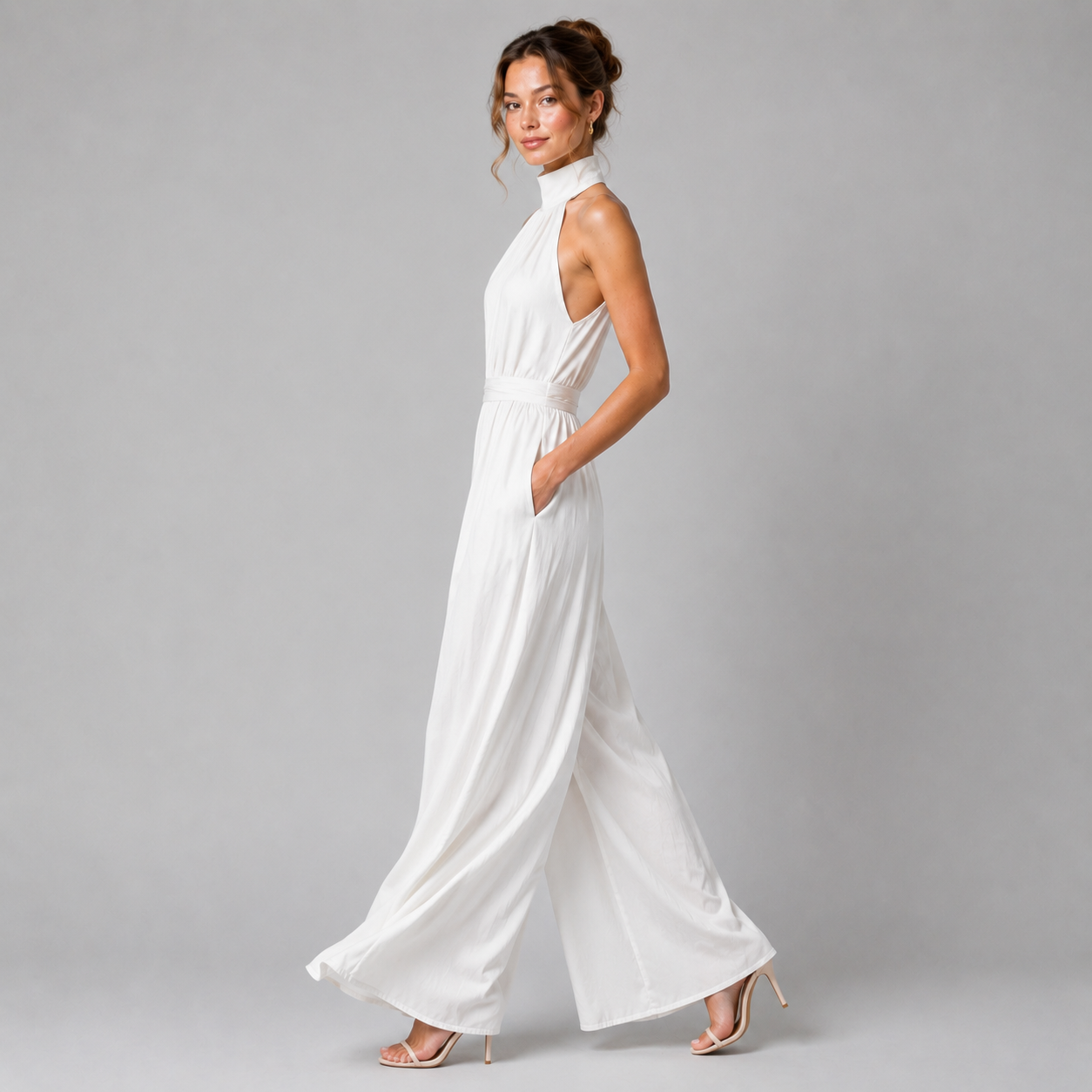 A woman modeling a white sleeveless high-neck wide-leg jumpsuit against a solid gray studio background.