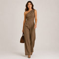 A woman models a brown one-shoulder wide-leg jumpsuit, paired with a matching brown handbag and brown heels, against a neutral light gray studio background.