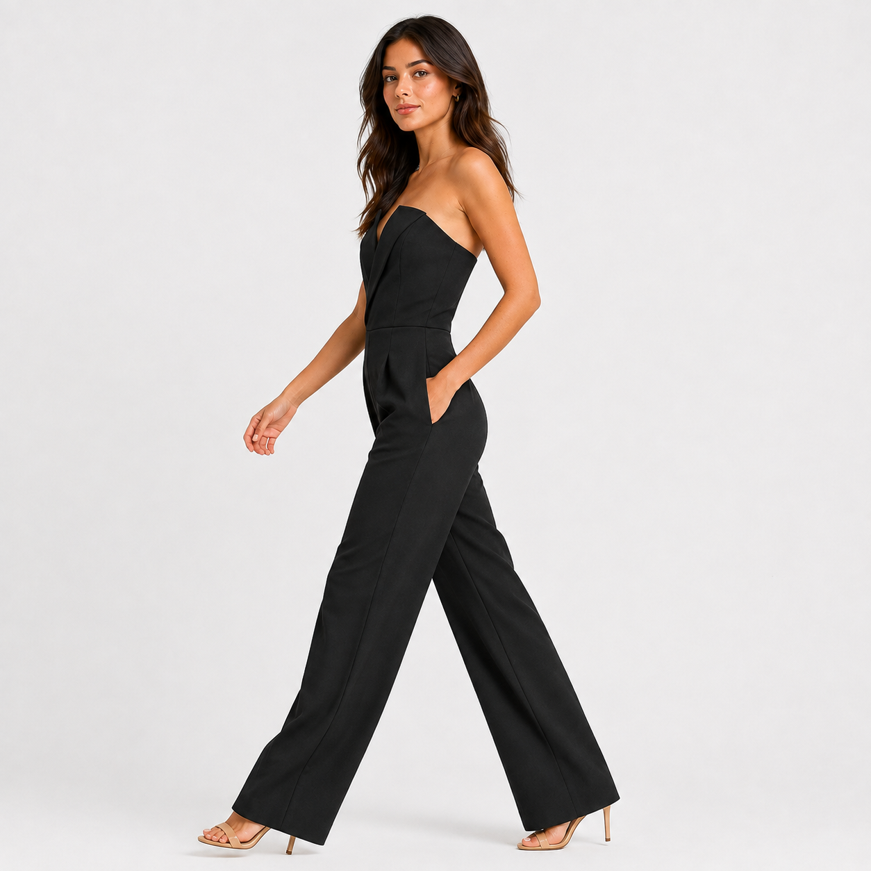 A woman modeling a black strapless jumpsuit with wide-leg pants and side pockets, paired with nude high-heeled sandals, against a light gray studio background.