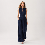 A woman modeling a sleeveless navy blue jumpsuit with vertical pleats, a keyhole neckline, and wide legs, standing against a plain light gray studio background.