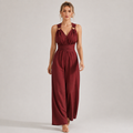 A woman modeling a sleeveless burgundy jumpsuit with a halter-style neckline, cutout details at the shoulders, a defined waist, and wide legs, photographed against a plain light gray studio background.