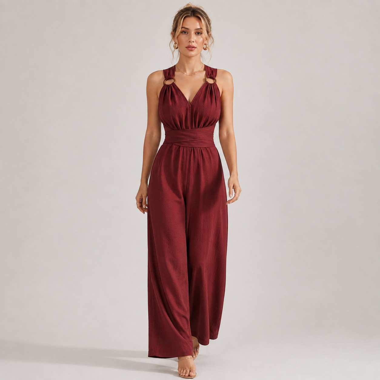 A woman modeling a sleeveless burgundy jumpsuit with a halter-style neckline, cutout details at the shoulders, a defined waist, and wide legs, photographed against a plain light gray studio background.