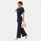 A woman models a navy blue two-piece jumpsuit with a tie waist and wide-leg pants, styled with a beige handbag and beige heels against a light gray background.