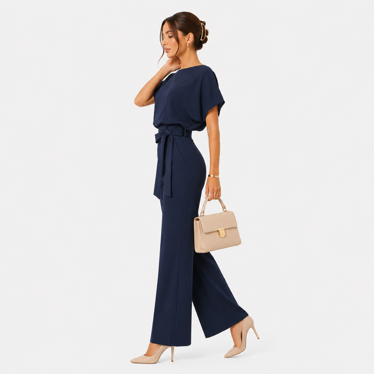 A woman models a navy blue two-piece jumpsuit with a tie waist and wide-leg pants, styled with a beige handbag and beige heels against a light gray background.