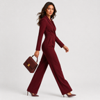A woman models a deep burgundy long-sleeve jumpsuit with a cinched waist and wide legs, holding a matching burgundy handbag with a gold clasp against a light gray studio background.