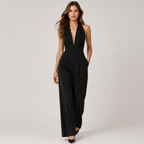 A woman modeling a black sleeveless halter-neck jumpsuit with a deep V-neckline and wide-leg pants, photographed against a neutral light gray studio background.