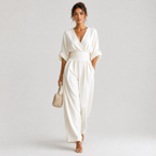 A woman models a white wrap-style jumpsuit with elbow-length sleeves, a defined waistband, and wide legs, holding a small beige handbag against a plain light gray studio background.