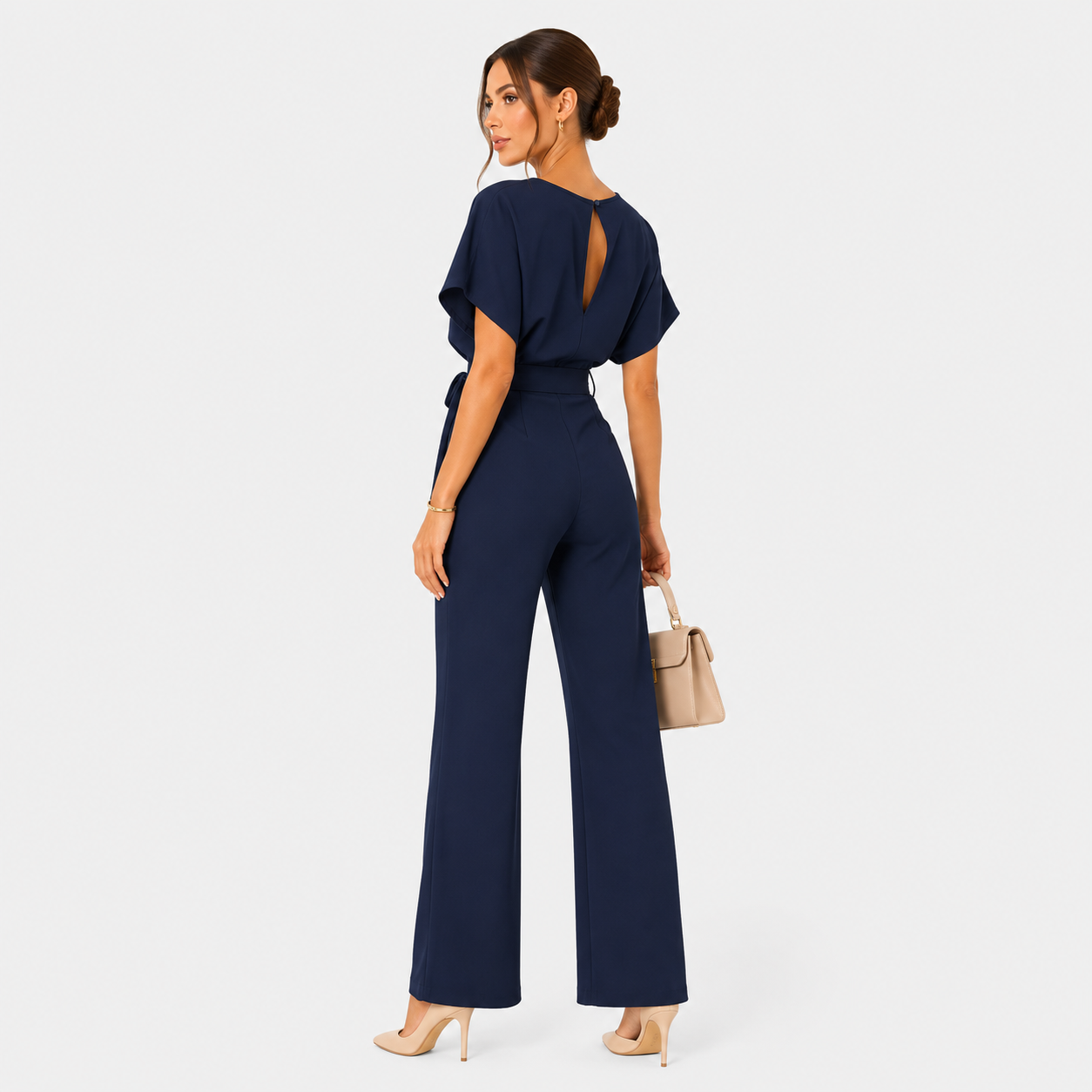 A woman modeling a navy blue jumpsuit from the back, featuring flutter sleeves, a keyhole back cutout, and wide-leg pants. She is holding a beige handbag and wearing beige high heels against a light gray studio background.