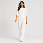 A woman modeling a white jumpsuit with elbow-length sleeves, a boat neckline, and decorative gold chain accents on the sleeves, photographed against a light gray studio background.