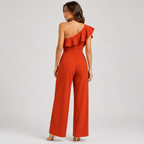 A woman modeling a bright rust-orange one-shoulder jumpsuit from the back, featuring a ruffled neckline and wide-leg pants, set against a plain light gray studio background.