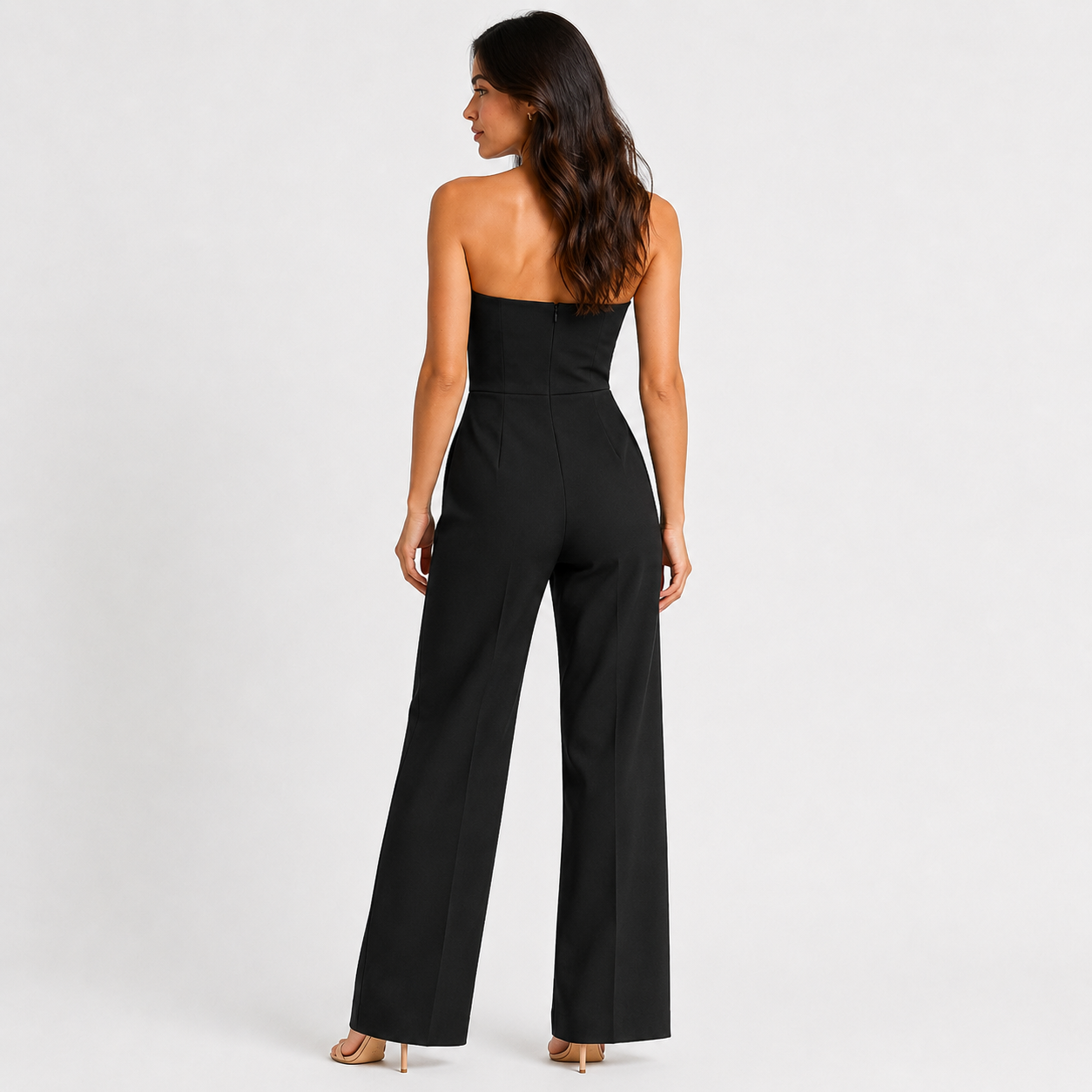 A woman viewed from the back wearing a black strapless wide-leg jumpsuit, standing against a plain light gray studio background.