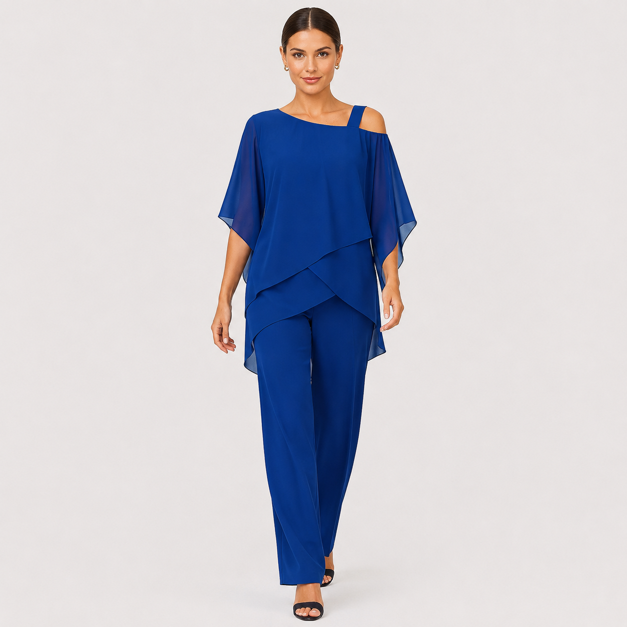 A woman modeling a royal blue two-piece outfit consisting of a draped cold-shoulder top and matching wide-leg pants, photographed against a light gray studio background.