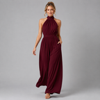 A woman modeling a sleeveless, high-neck, floor-length burgundy maxi dress with a gathered waist and side pockets, photographed against a solid gray studio background.