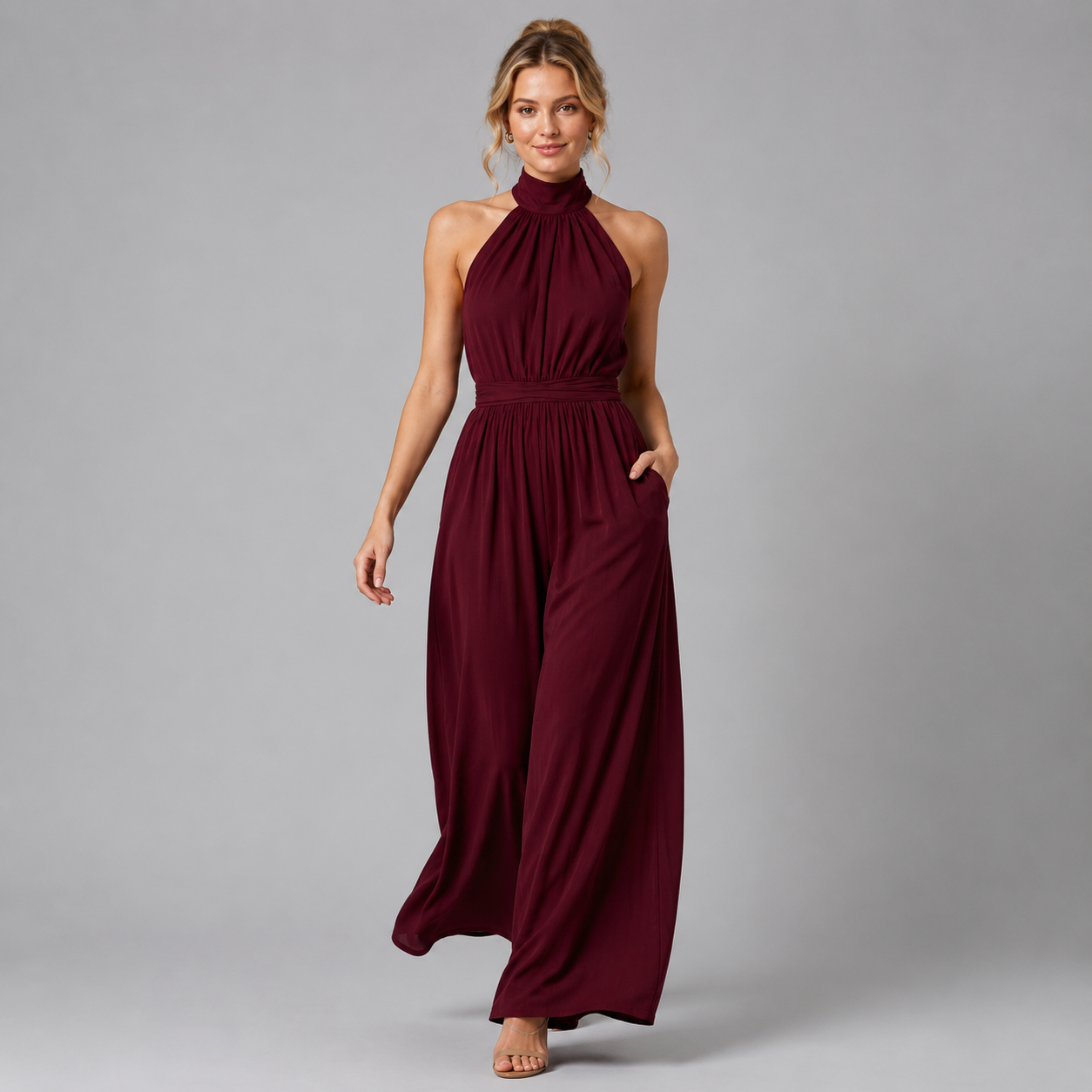 A woman modeling a sleeveless, high-neck, floor-length burgundy maxi dress with a gathered waist and side pockets, photographed against a solid gray studio background.