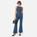 A woman modeling a sleeveless blue denim jumpsuit with a front zipper, flared legs, and side cutouts, styled with black sunglasses, a small black handbag, and black heels against a light gray studio background.