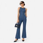 A woman modeling a sleeveless blue denim jumpsuit with a front zipper, flared legs, and side cutouts, styled with black sunglasses, a small black handbag, and black heels against a light gray studio background.