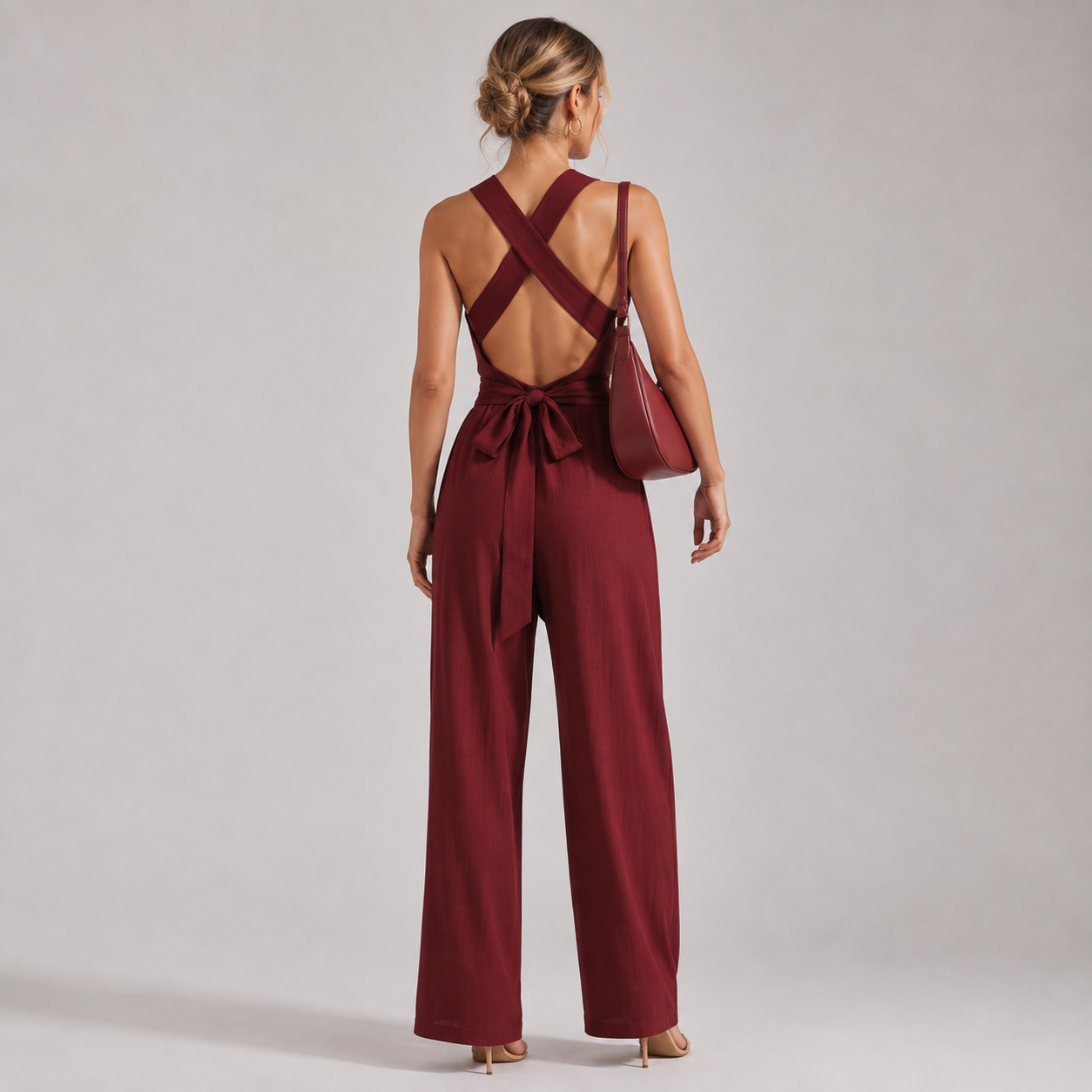 A woman viewed from behind wearing a burgundy wide-leg jumpsuit with crisscross back straps and a tie at the waist, standing against a light gray studio background and carrying a matching shoulder bag.