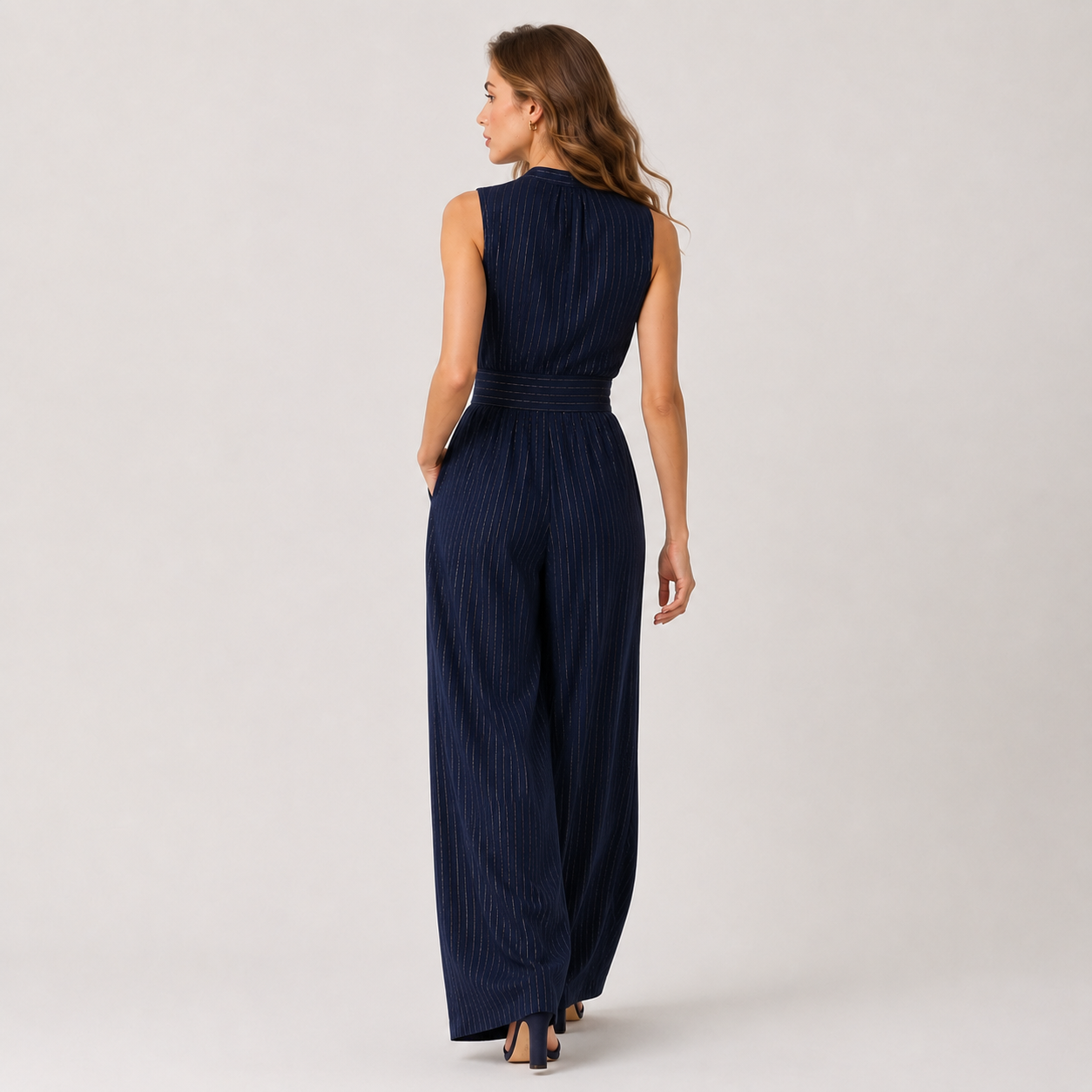 A woman wearing a navy blue sleeveless jumpsuit with vertical pleats, shown from the back against a plain light gray studio background.